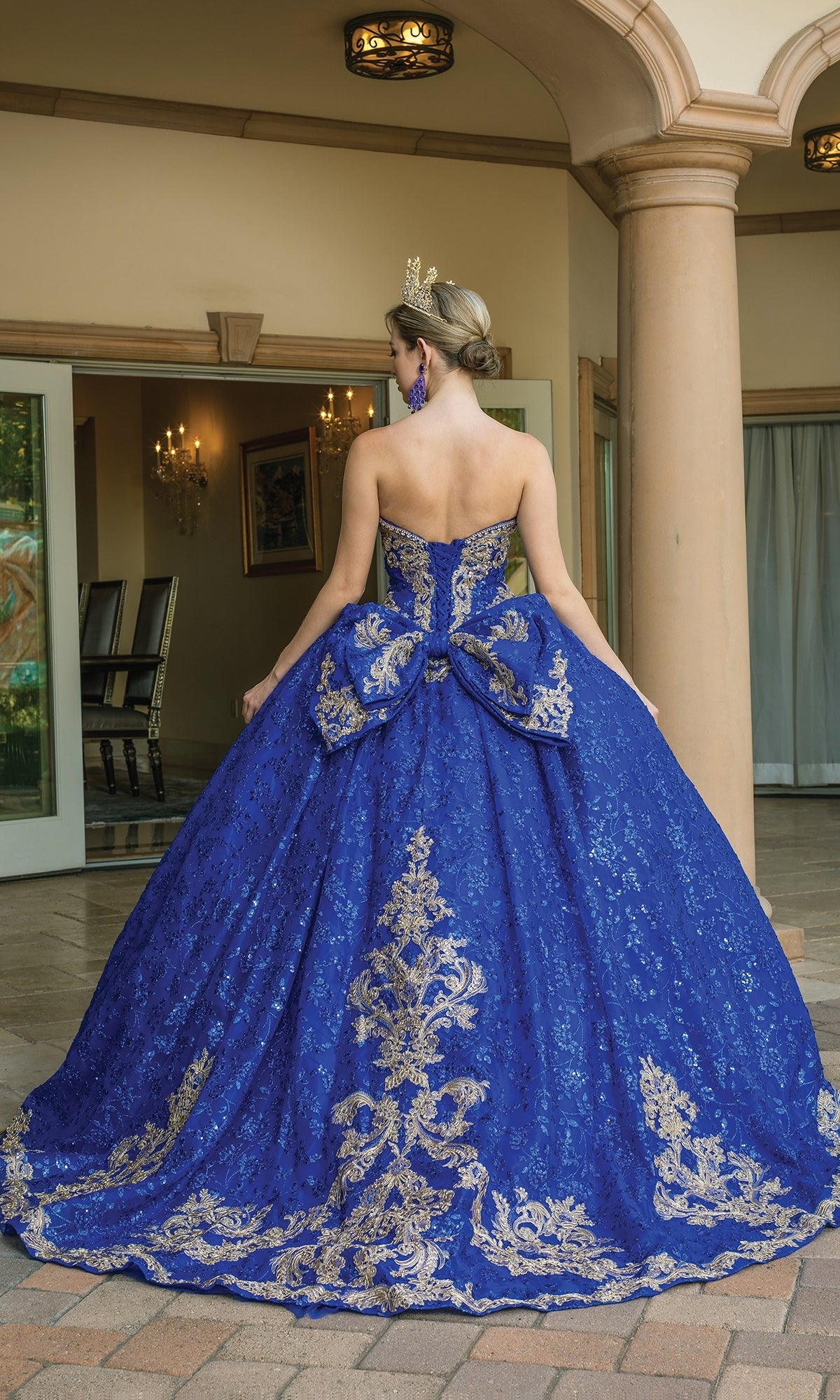 A woman wearing the Quinceanera Dress 1650 By Dancing Queen, a strapless ball gown with gold lace embroidery and a large bow, stands outside near columns with her back to the camera.